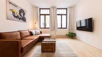 Una sala de estar con un sofá de cuero marrón, una mesa de centro de madera, un televisor montado en la pared y una ventana con cortinas.