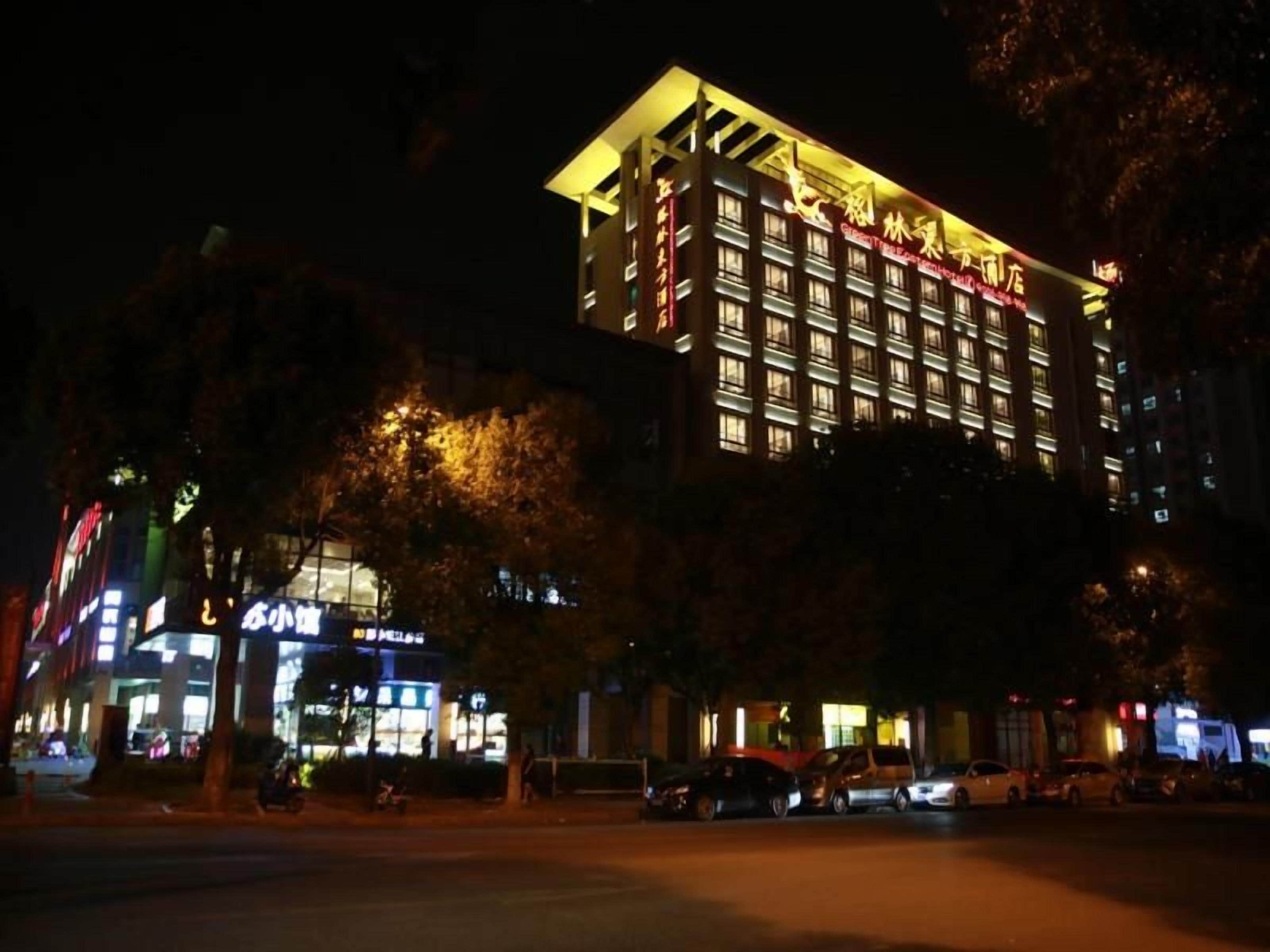 A multi-story hotel with illuminated signage, a parking area in front, and a tree-lined street.