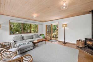 A modern living room with a grey sofa, wooden coffee table, wicker chairs, a fireplace, and large windows offering a view of greenery. - Atwood (Aireys Inlet)