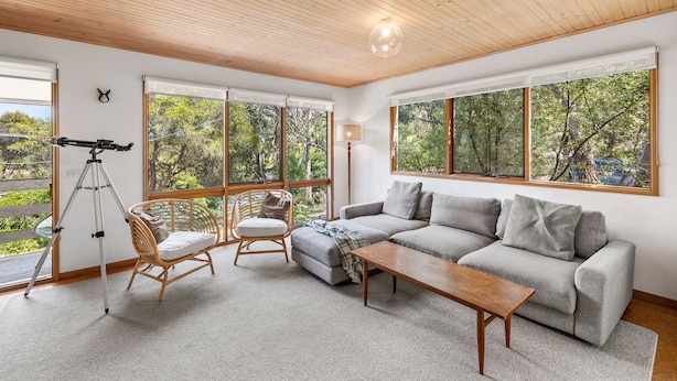 A spacious living room with a large window, a sofa, a wooden coffee table, a wicker chair, and a telescope.