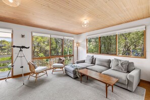 Ein geräumiges Wohnzimmer mit großem Fenster, einer Couch, einem Holztisch, einem Korbstuhl und einem Teleskop.