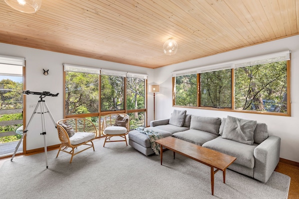 A spacious living room with a large window, a sofa, a wooden coffee table, a wicker chair, and a telescope.