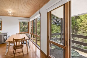 A dining area with a wooden table and chairs, large windows, and a view of greenery outside.