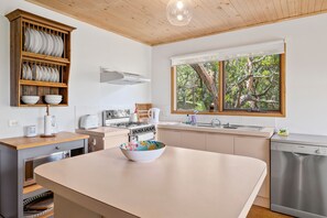 A modern kitchen with a central island, wooden cabinetry, a built-in dishwasher, and a window with a view of trees.