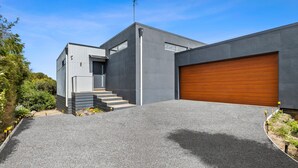 Ein modernes, zweigeschossiges Haus mit einer grauen Fassade, einer Holztür zur Garage und einer Treppe zum Eingang.