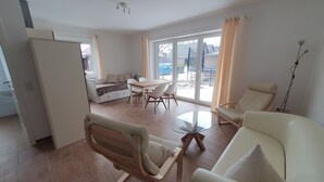 A modern living room with a sofa, a dining table, and a reclining chair. There is a small kitchen area with a sink and a window. - Haus Taybeerweg (Aurich)