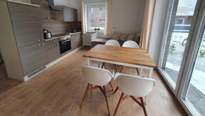 A modern kitchen with wooden cabinets, a dining table with chairs, and a large window offering a view of the outside. - Haus Taybeerweg (Aurich)