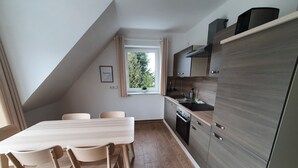 A modern kitchen with a built-in oven, a sink, and wooden cabinets. There is a dining table with chairs and a window with curtains. - Haus Taybeerweg (Aurich)