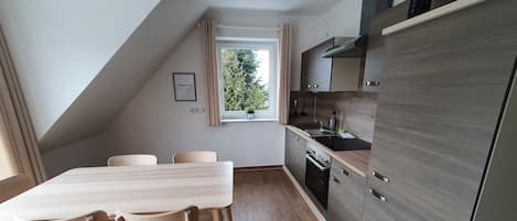 A modern kitchen with a built-in oven, a sink, and wooden cabinets. There is a dining table with chairs and a window with curtains.