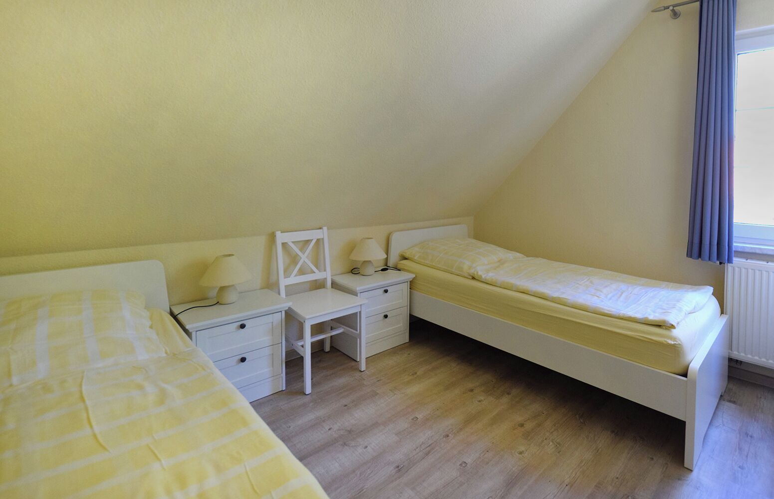 A small, single-room bedroom with a sloped ceiling, a desk, two beds, and a radiator.