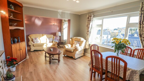 A spacious living room with a wooden floor, a sofa set, a dining table with chairs, and a view of the city through large windows. - Lavender Cottage (Castletownbere)