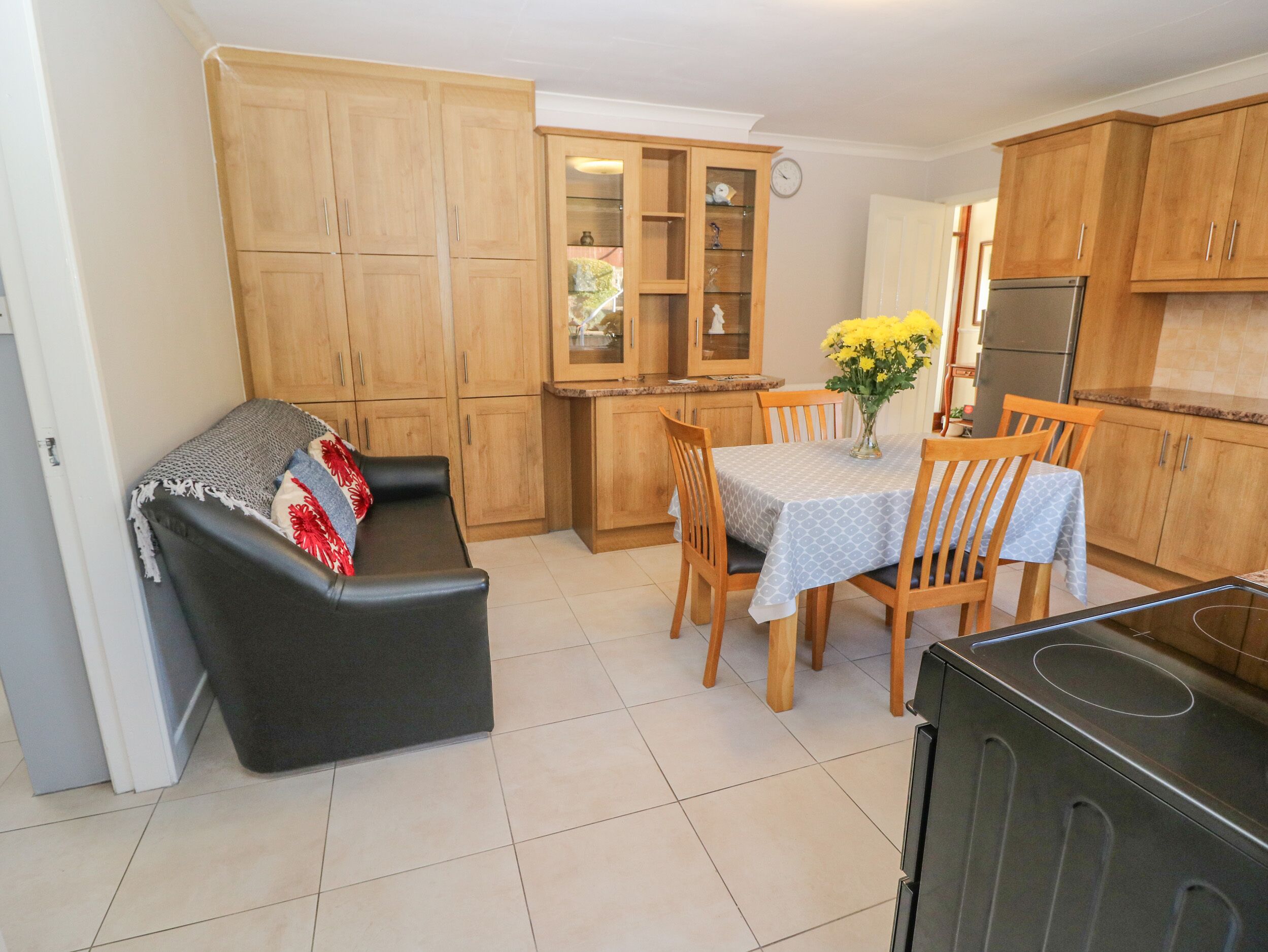 A compact kitchen with wooden cabinets, a dining table with chairs, a black sofa, and a refrigerator.