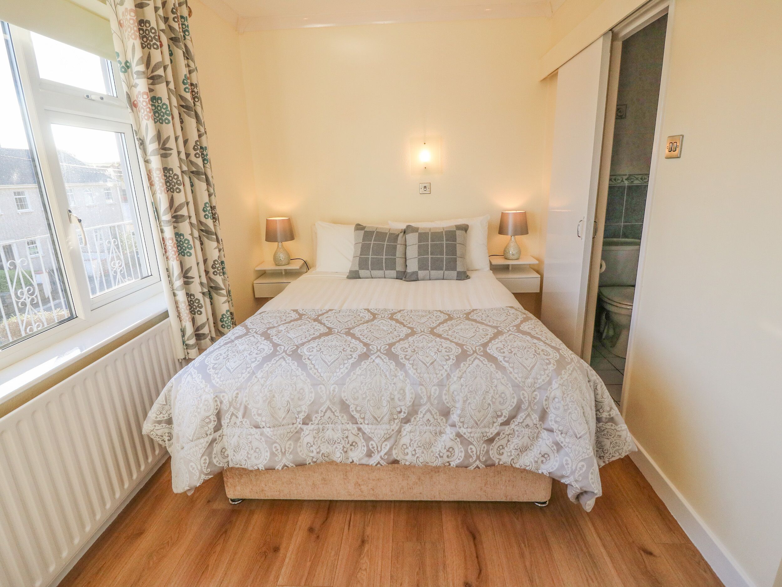 Une chambre avec un grand lit, deux tables de chevet avec des lampes, un radiateur, une fenêtre avec des rideaux et une vue sur l’extérieur.