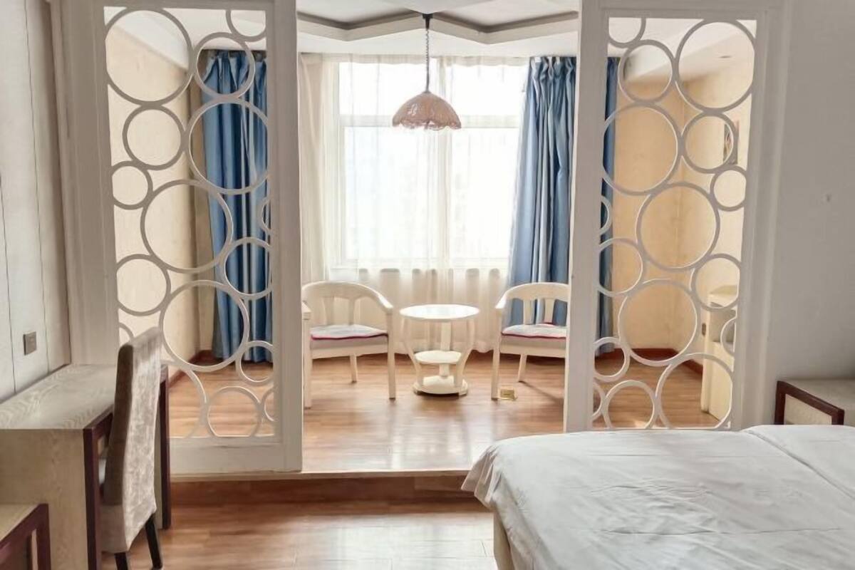 A bedroom with a bed, a dining table with chairs, and a view of the outside through a window with curtains.