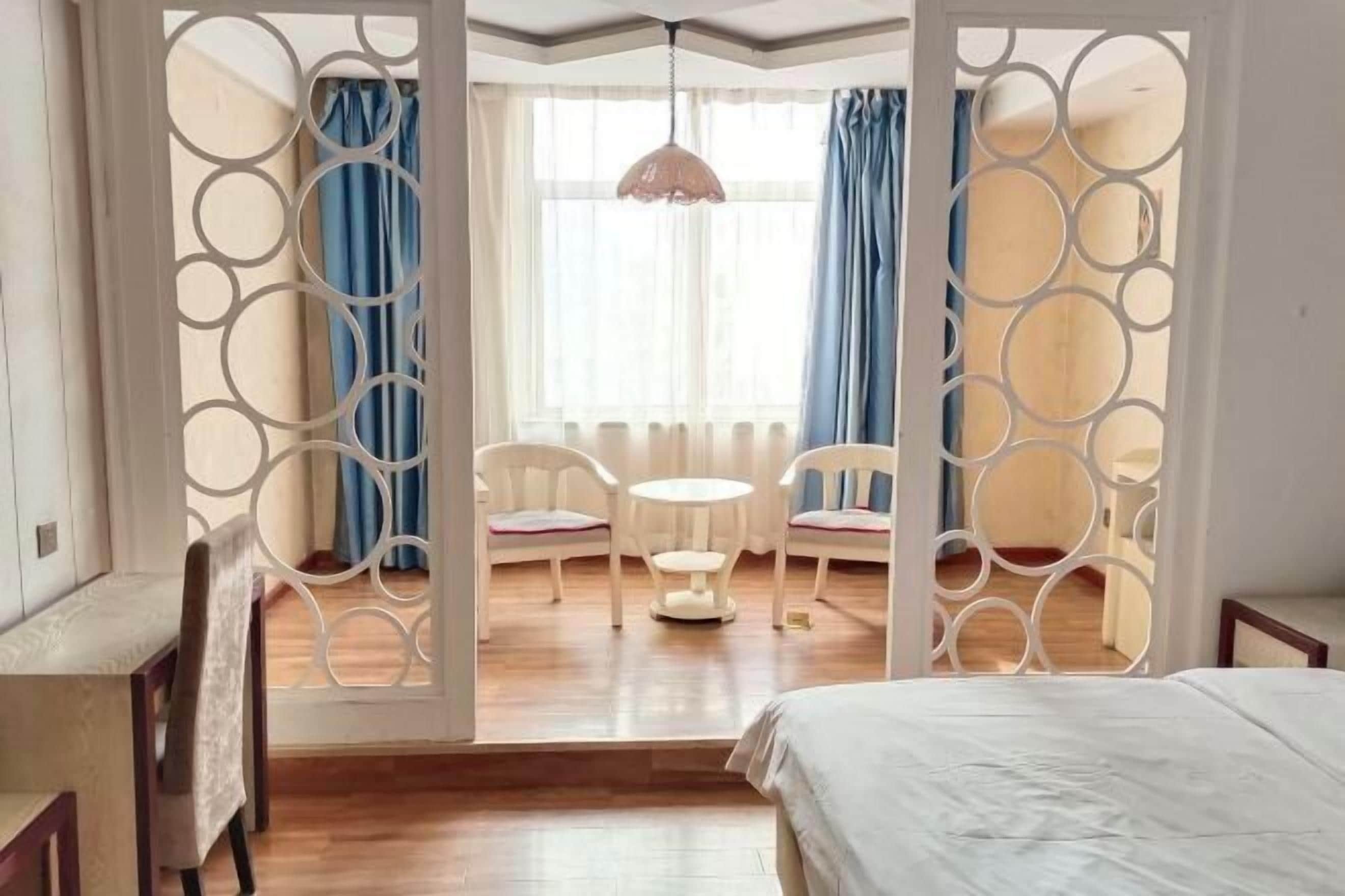 Un dormitorio con una cama, un escritorio y una silla. Se ve una ventana con cortinas y una lámpara colgante.
