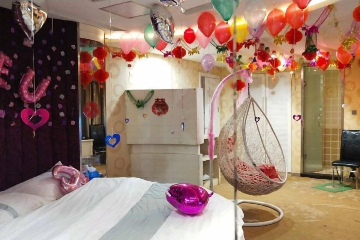 A hotel room with a bed, a hanging chair, and a wall decorated with balloons and heart-shaped items.