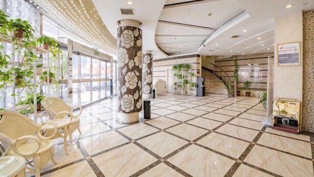 Um hall amplo com piso de azulejos, um pilar central com design floral, cadeiras de vime e plantas em vasos.