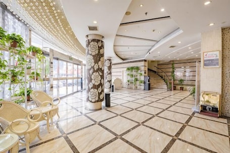 A spacious lobby with a tiled floor, a central column with floral design, wicker chairs, and potted plants.. Mingdu Holiday Hotel