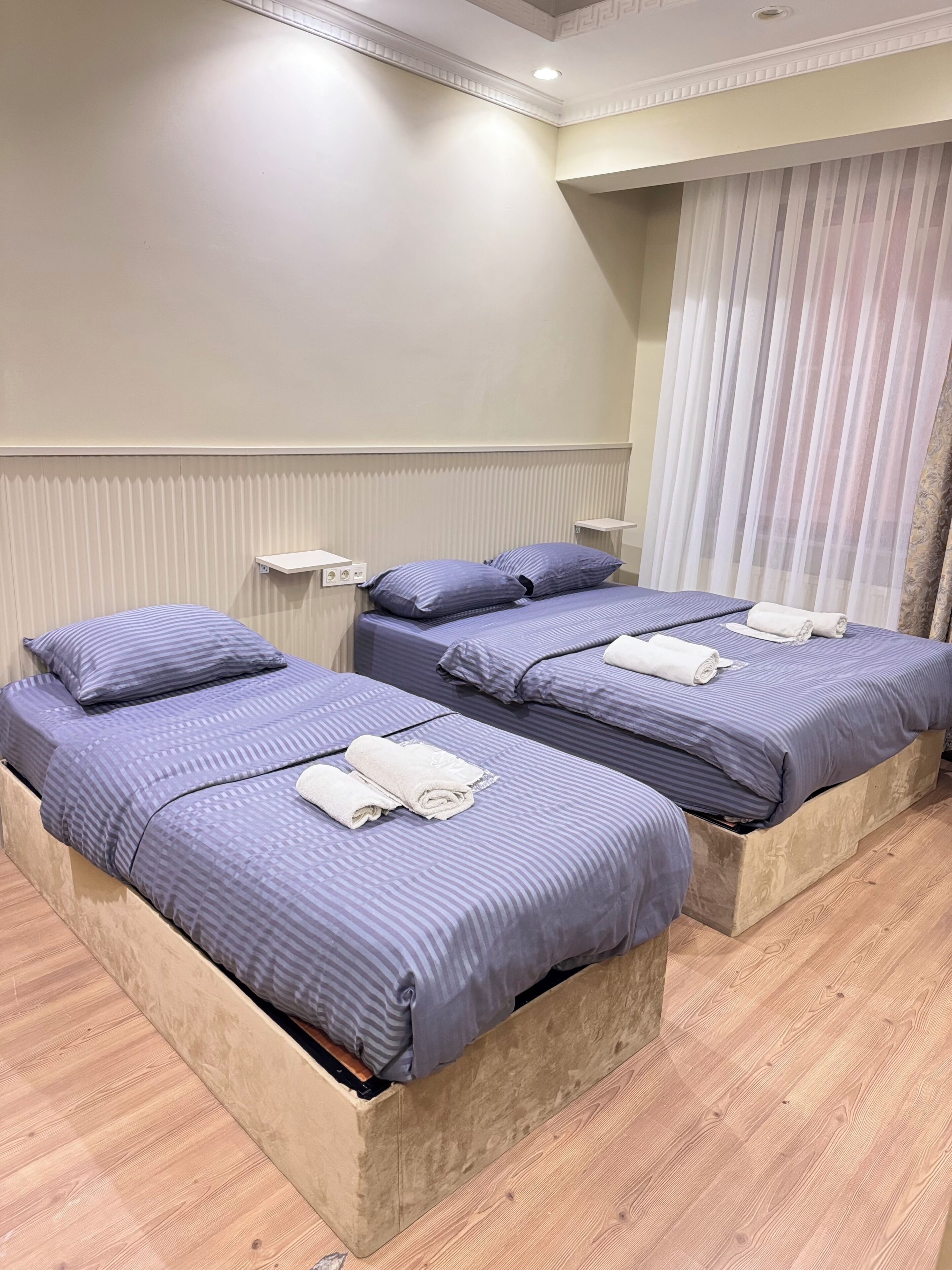 Una habitació d'hotel amb tres llits, cadascun amb una tovallola enrotllada. La habitació té un paviment de fusta i una paret blanca amb una obertura de ventilació.