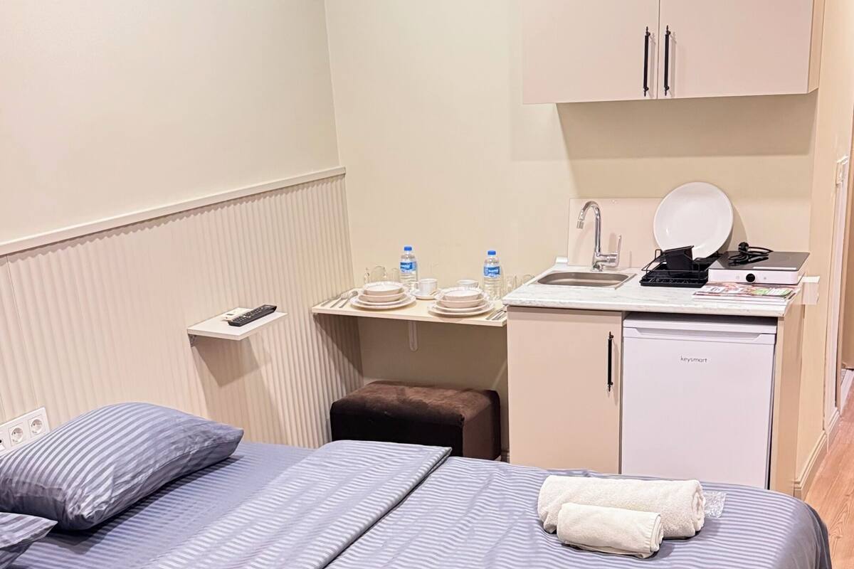 A hotel room with a bed, a desk with a kettle, a kettle, a small fridge, and a cabinet.