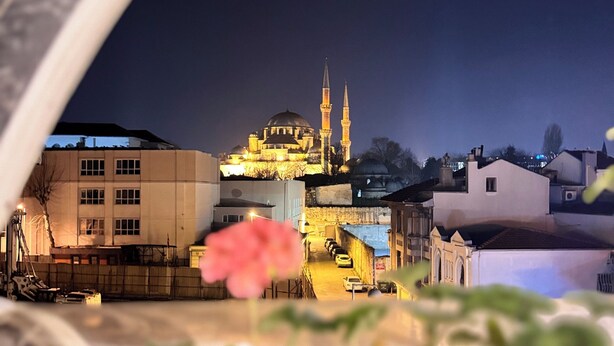 Vista nocturna de un paisaje urbano con una mezquita destacada iluminada contra el cielo oscuro, rodeada de edificios y una flor rosada en primer plano.