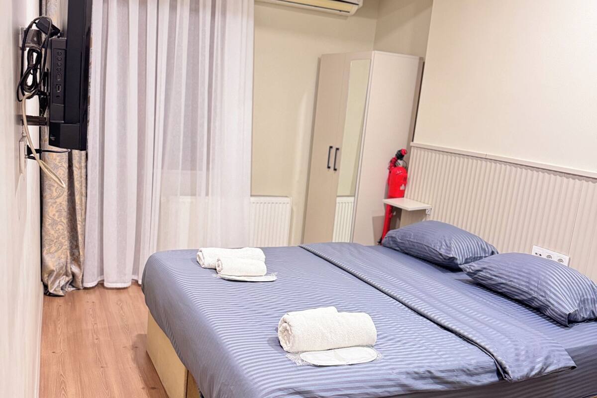 A bedroom with a bed, curtains, an air conditioning unit, a wardrobe, a mirror, and a television.