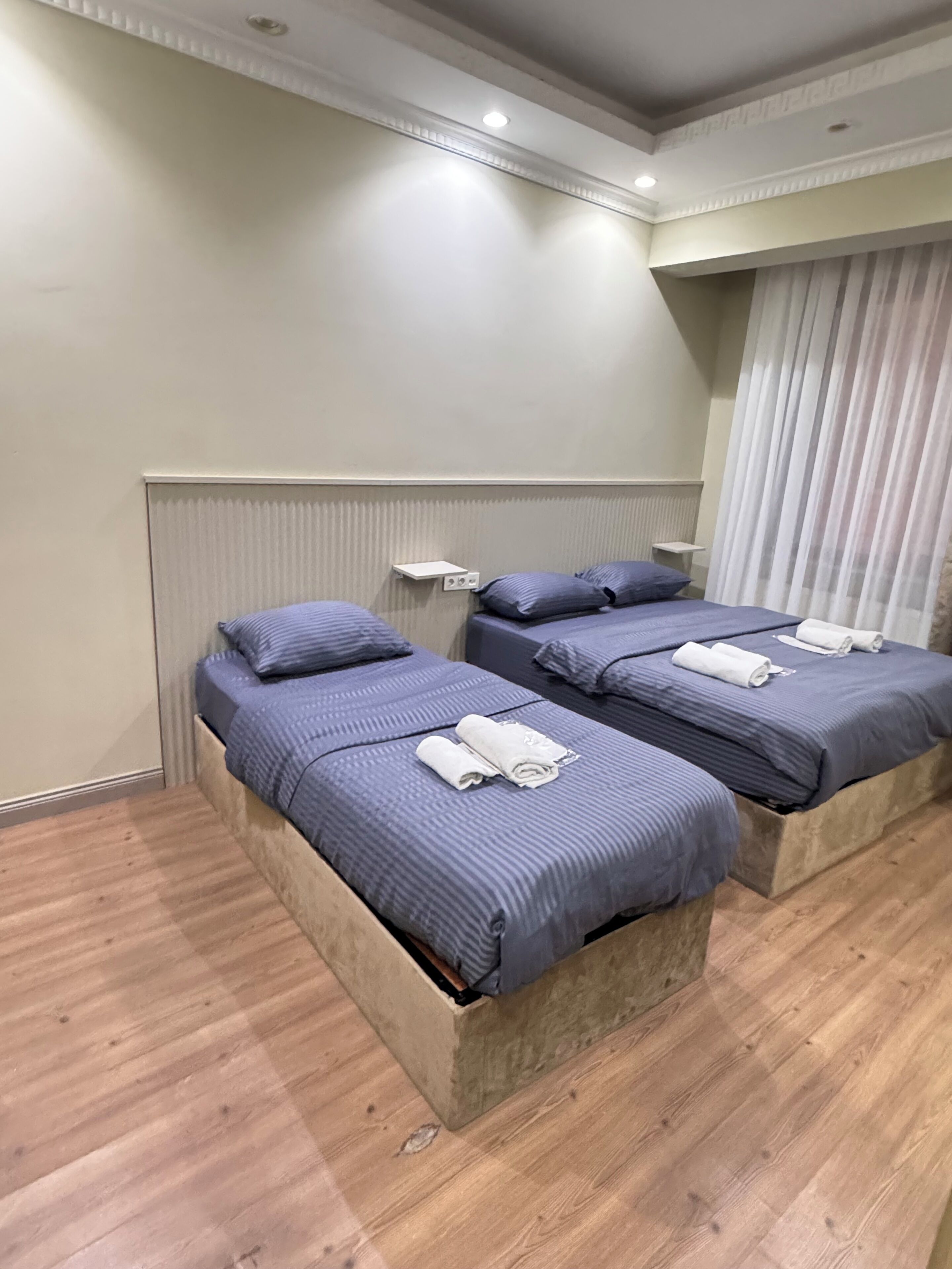 Quarto de hotel com duas camas, piso de madeira, radiador e janela com cortinas.