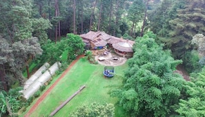 Um chalé isolado, rodeado por floresta densa, com um pátio circular, um trampolim, uma piscina pequena e uma construção com telhado de palha.