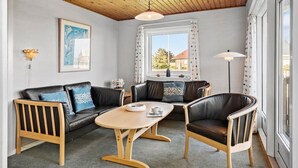 Un salon chaleureux avec un plafond en bois, un canapé en cuir noir, une table basse en bois et une vue sur l’extérieur par une fenêtre.
