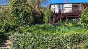 A rustic cabin with a wooden staircase leading to a deck, surrounded by lush greenery and flowering plants. - Treetops, a relaxing haven in Bantham South Devon (Bantham)