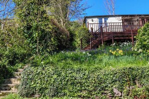 A rustic cabin with a wooden staircase leading to a deck, surrounded by lush greenery and flowering plants. - Treetops, a relaxing haven in Bantham South Devon (Bantham)