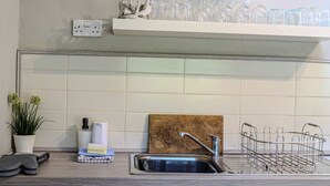A kitchen with a sink, a wooden cutting board, a metal dish rack, a potted plant, and a shelf with glassware. - Treetops, a relaxing haven in Bantham South Devon (Bantham)
