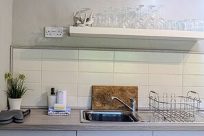 A kitchen with a sink, a wooden cutting board, a metal dish rack, a potted plant, and a shelf with glassware. - Treetops, a relaxing haven in Bantham South Devon (Bantham)