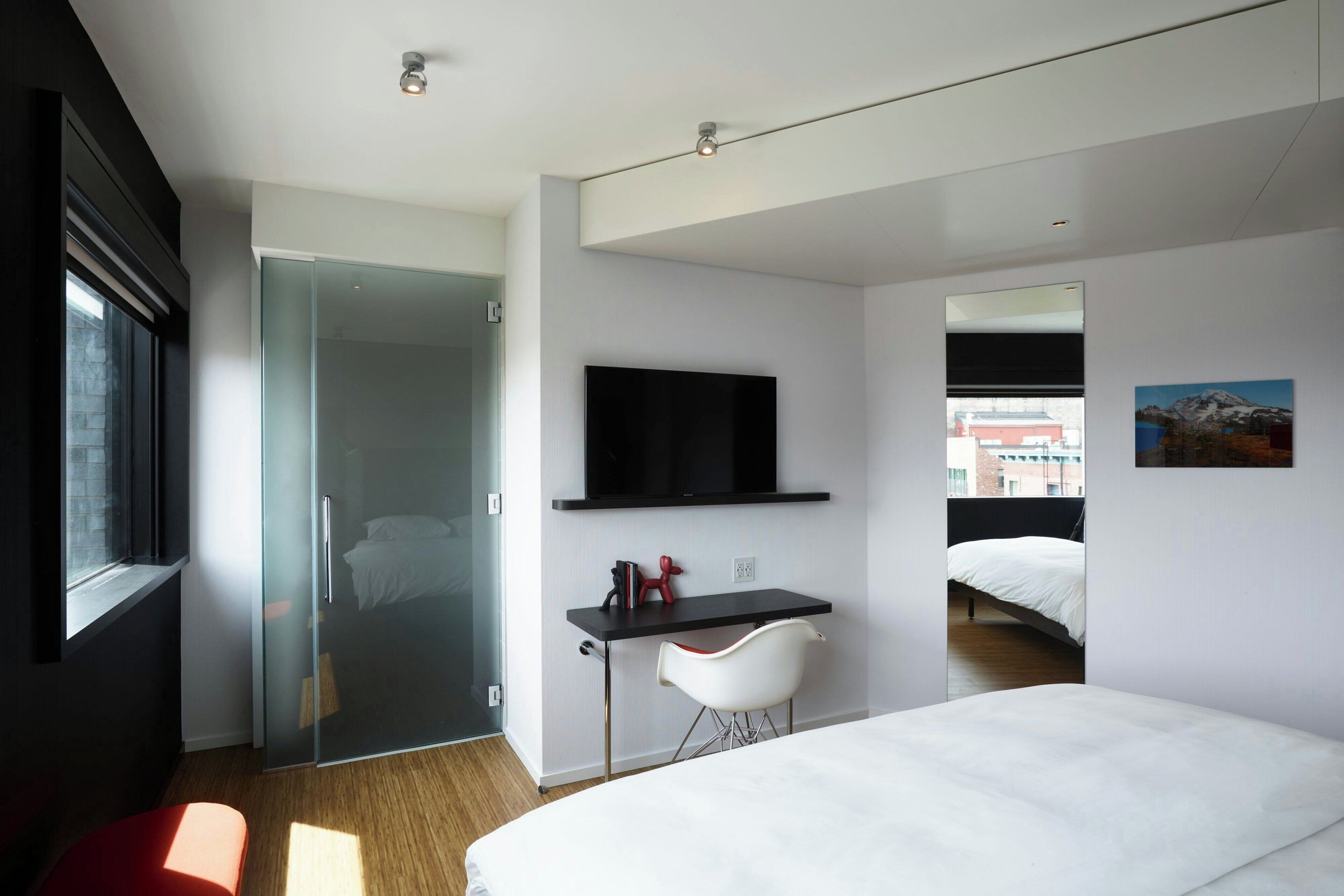 Une chambre d’hôtel moderne équipée d’un lit, d’un bureau avec une chaise, d’une télévision et d’une porte en verre donnant sur une salle de bain.