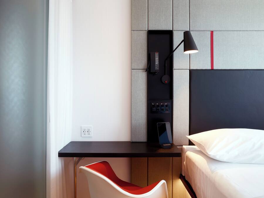 A modern hotel room with a desk, a red chair, a bed, and a wall-mounted lamp.