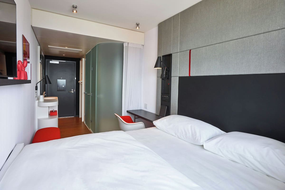 A modern hotel room with two beds, a desk, a chair, and a small kitchenette.
