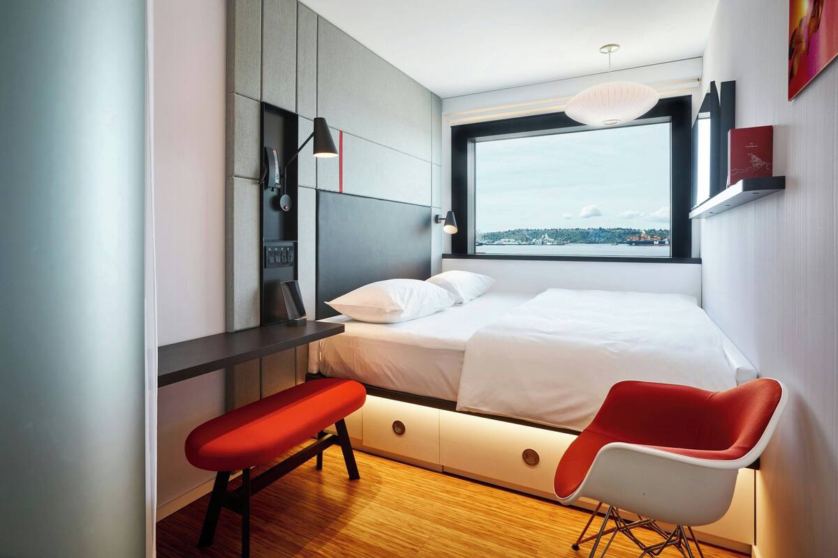 A modern bedroom with a bed, a bench, a desk, and a large window offering a city view.