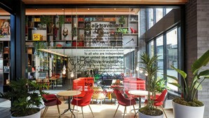 A modern interior with a glass wall displaying a welcoming message to travelers, shelves with books and decorative items, and a seating area with red chairs and potted plants. - citizenM Seattle Pioneer Square (Seattle)