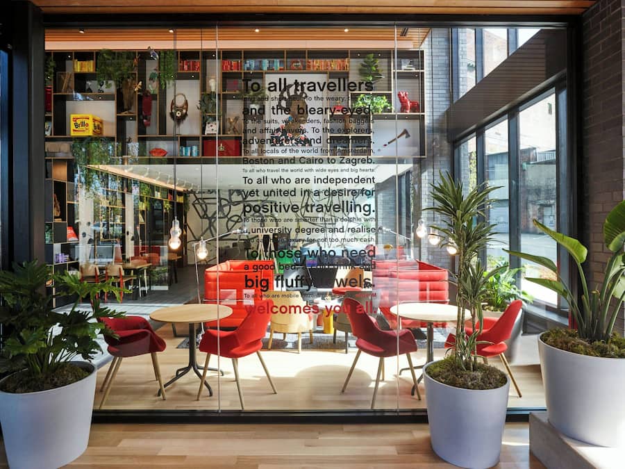 A modern interior with a glass wall displaying a welcoming message to travelers, shelves with books and decorative items, and a seating area with red chairs and potted plants.