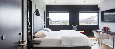 A modern bedroom with a large bed, a desk, and a view of a cityscape through large windows.