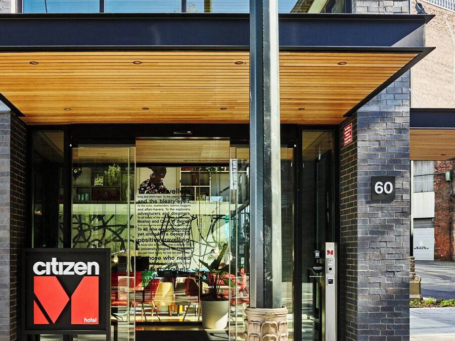 Una entrada de hotel moderna con fachada de vidrio, un alero de madera y un llamativo letrero que dice 'Citizen Hotel'.