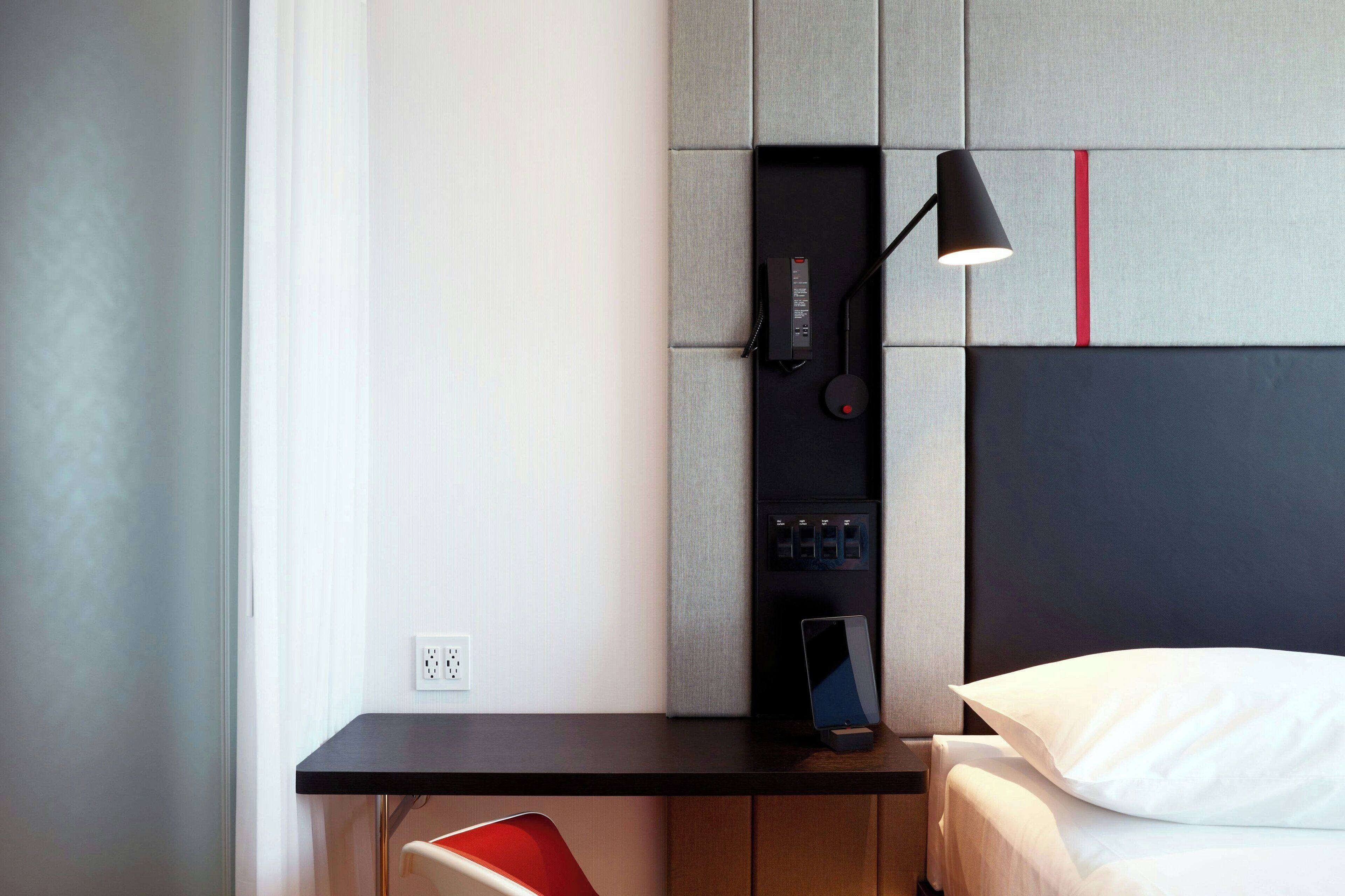 Una habitación de hotel moderna con escritorio, cama, una silla roja y un panel de control en la pared.