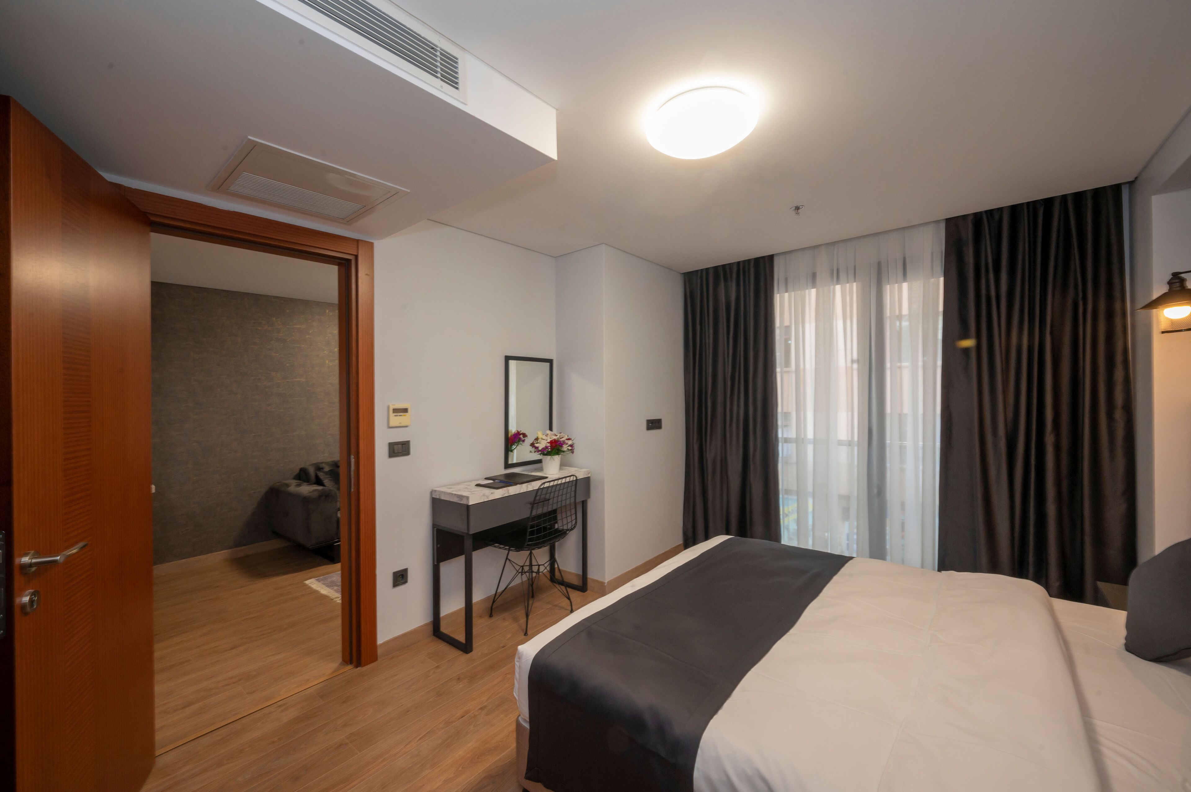 A modern hotel room with a large bed, a desk with a chair, a mirror, and a window with curtains.