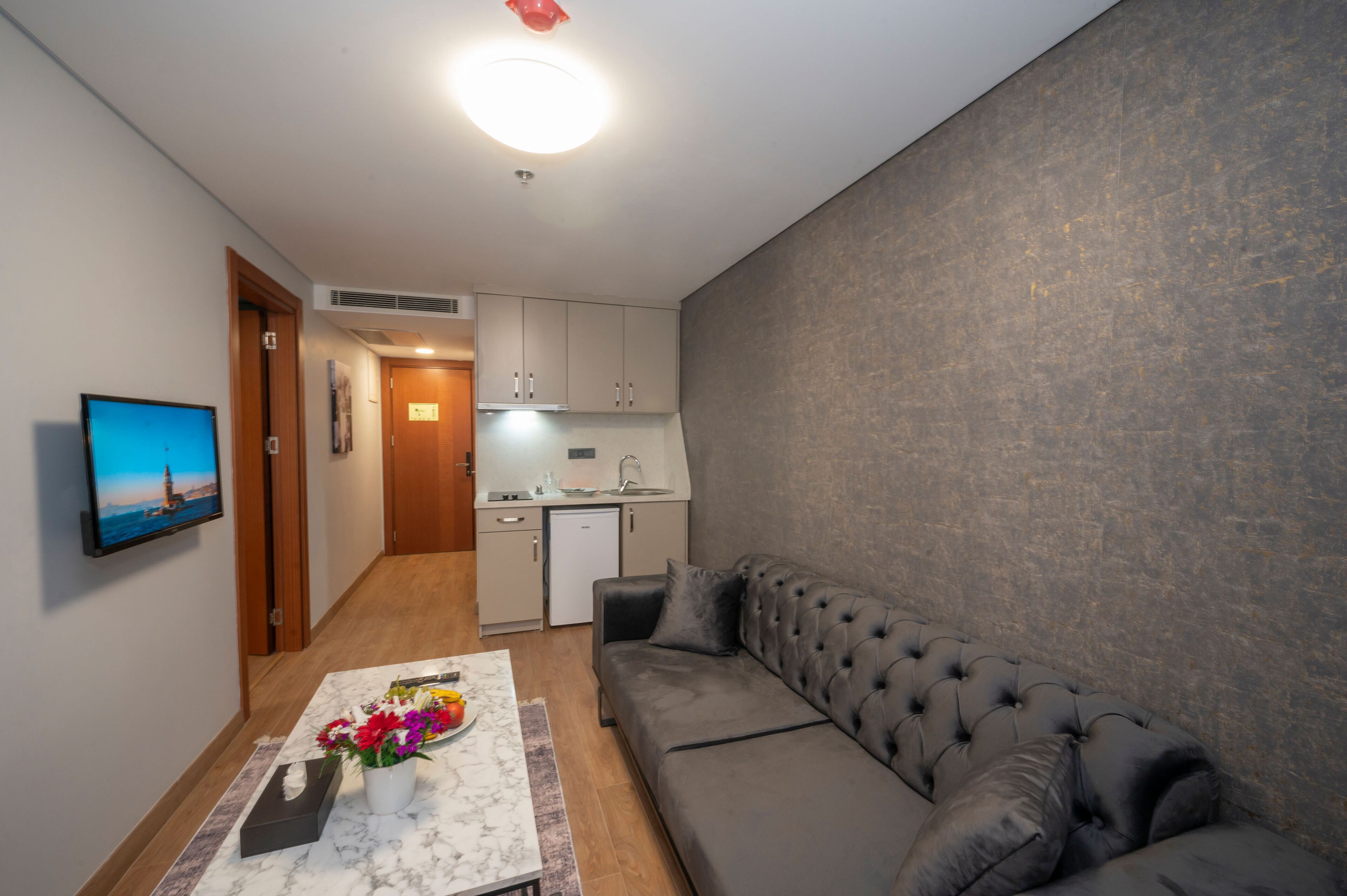 A compact living space with a sofa, a small table with flowers, a kitchen area, and a TV on the wall.