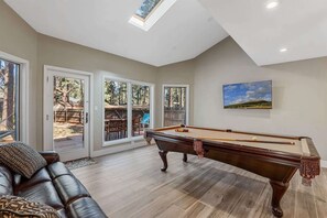 A spacious living room with a pool table, a comfortable leather couch, and a large window overlooking a backyard.