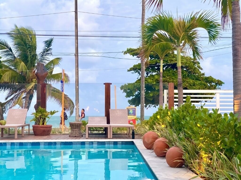 A pool area with lounge chairs, a view of the ocean, and palm trees.