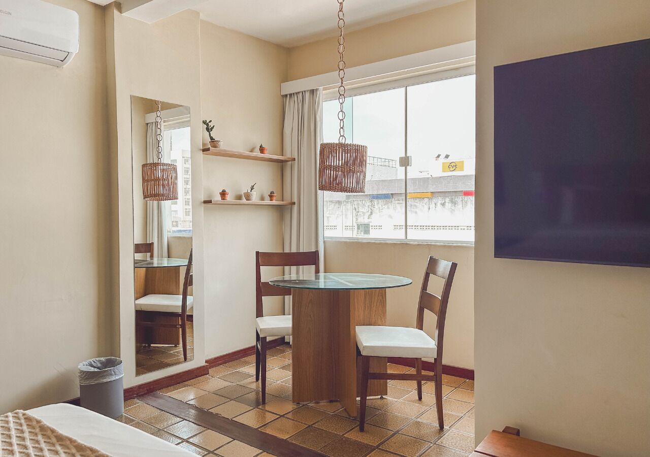 Una stanza compatta con un tavolo rotondo e quattro sedie, un piccolo ripiano con oggetti decorativi, un lampadario in vimini appeso e un'ampia finestra con vista sugli edifici.
