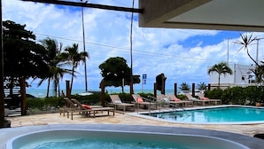 A pool area with lounge chairs and a hot tub, overlooking a beach with palm trees and a clear sky.