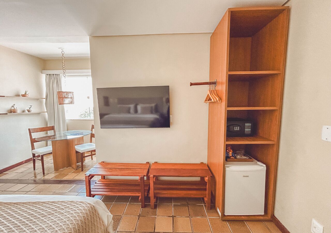A hotel room with a bed, a wooden bench, a small table with chairs, a television, a wooden shelf with a microwave, and a refrigerator.