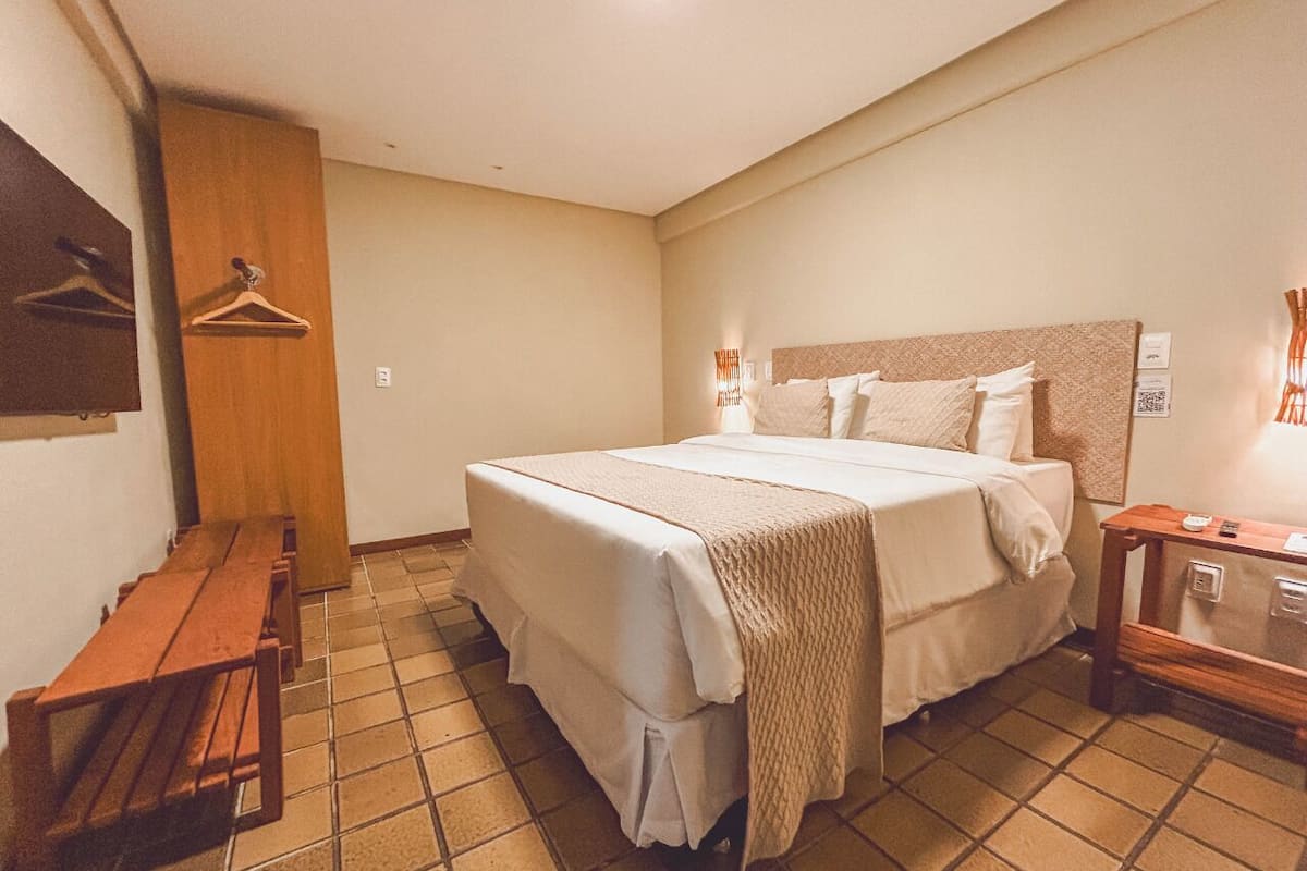 Una habitación de hotel con una cama, un banco, una percha de madera y una mesita de noche.